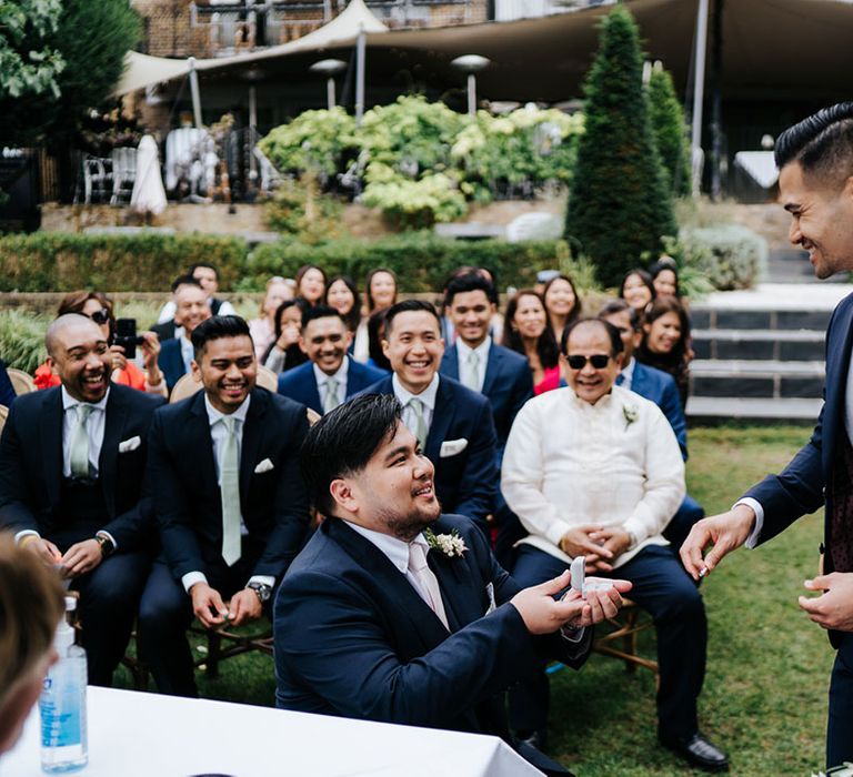 Ecstatic Groom stands in navy suit with a beautiful blush pink boutonniere smiling in stunning outdoor ceremony