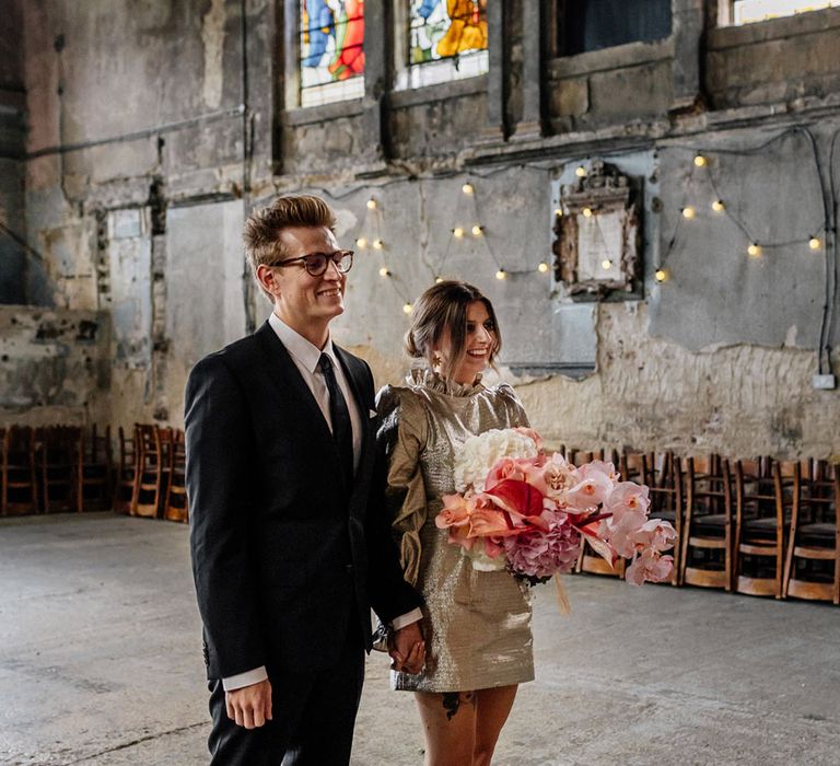 Edgy Bride in croc print short gold wedding dress dress and Groom in dark suit stand with colourful church windows surrounding them The Asylum London