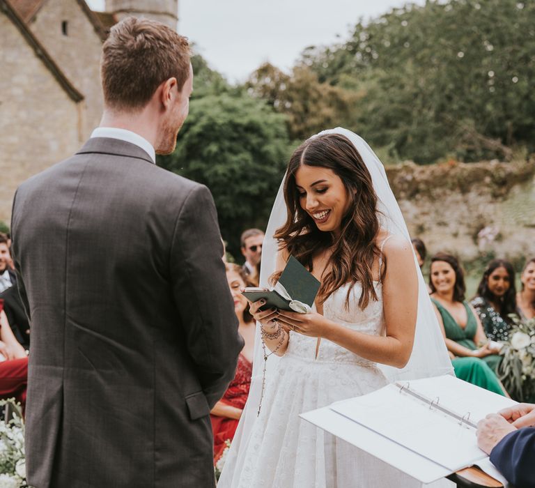 Bride in Pronovias wedding dress and veil reads vows during outdoor wedding ceremony at Notley Abbey as she stands opposite groom in grey suit 