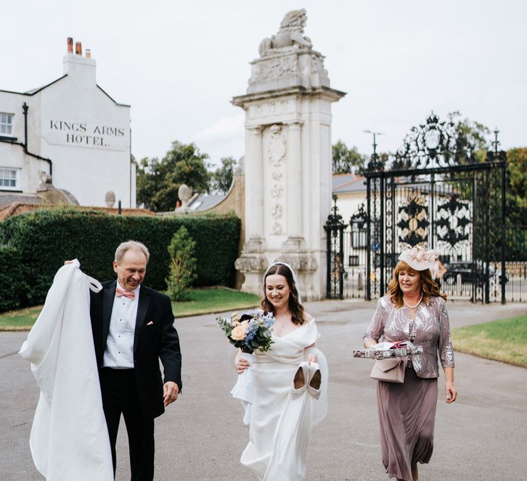 Bride walks with her parents on her wedding day as she wears flat shoes and carries her heels