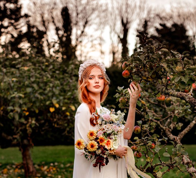 Bride in white wedding dress and cape with netted headpiece holds colourful pastel bridal bouquet and apple as she stands in apple orchard