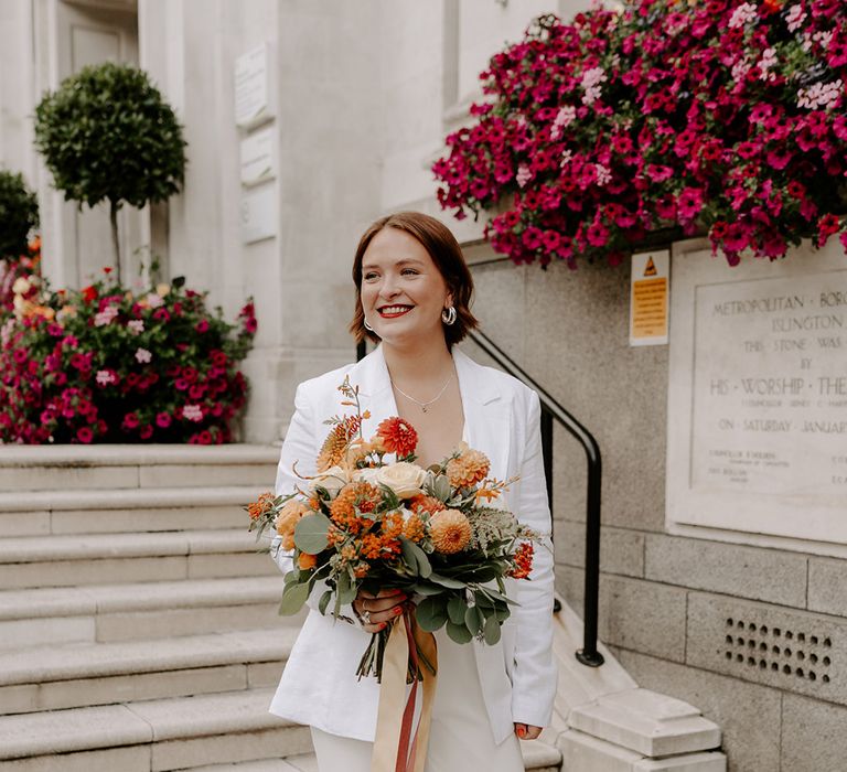 Bride in a jumpsuit holding an peach and orange flower bouquet at Islington Town Hall 