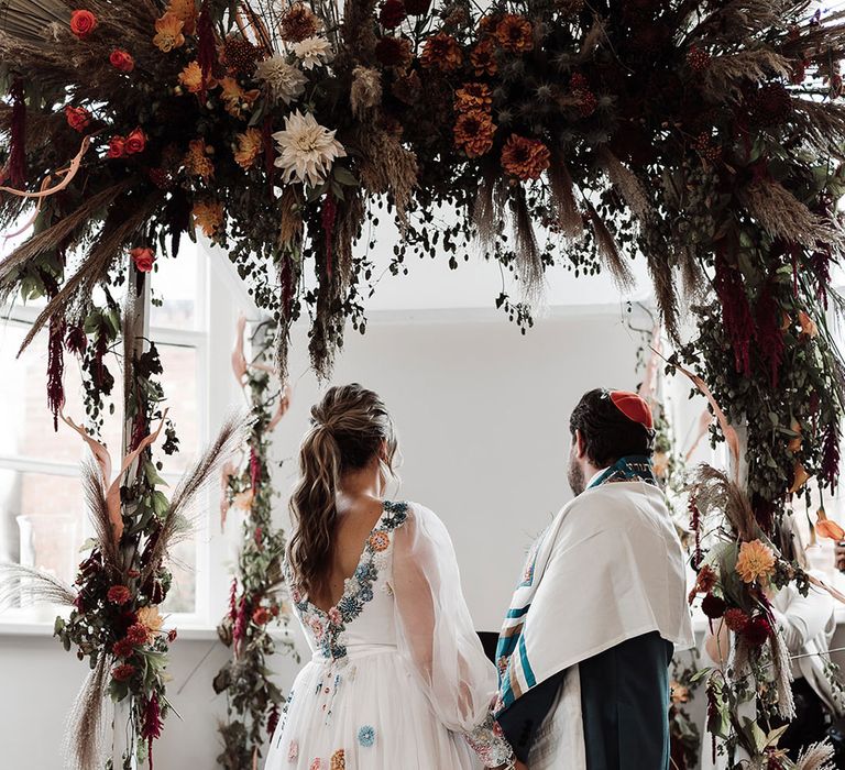 Bride in low back embroidered Victoria Sanders wedding dress with train stands with groom in green suit and red kippah under wedding arch with dried flower cloud 