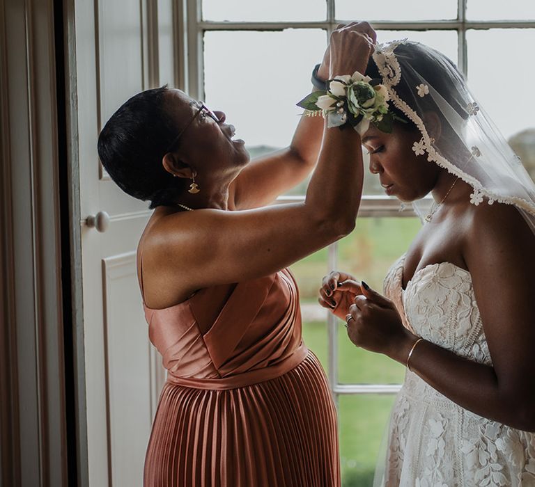 Mother of the bride in silk pleated dress helps bride put on her wedding veil with lace edge detail