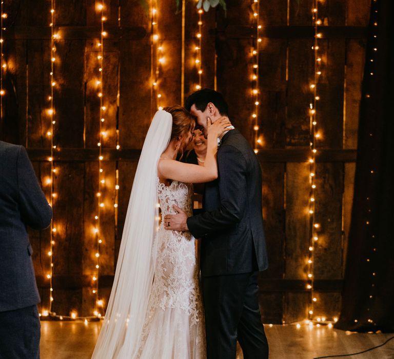 Bride in chapel length veil and lace wedding dress hugs groom in dark suit during fairy-lit wedding ceremony in barn