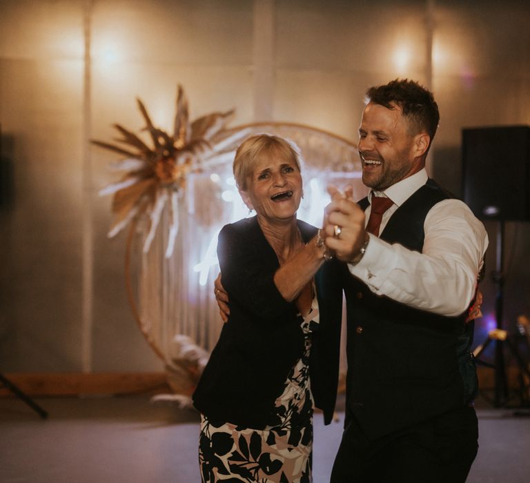 Groom in white shirt and waistcoat dances with wedding guests in patterned dress by neon wedding sign at barn wedding reception