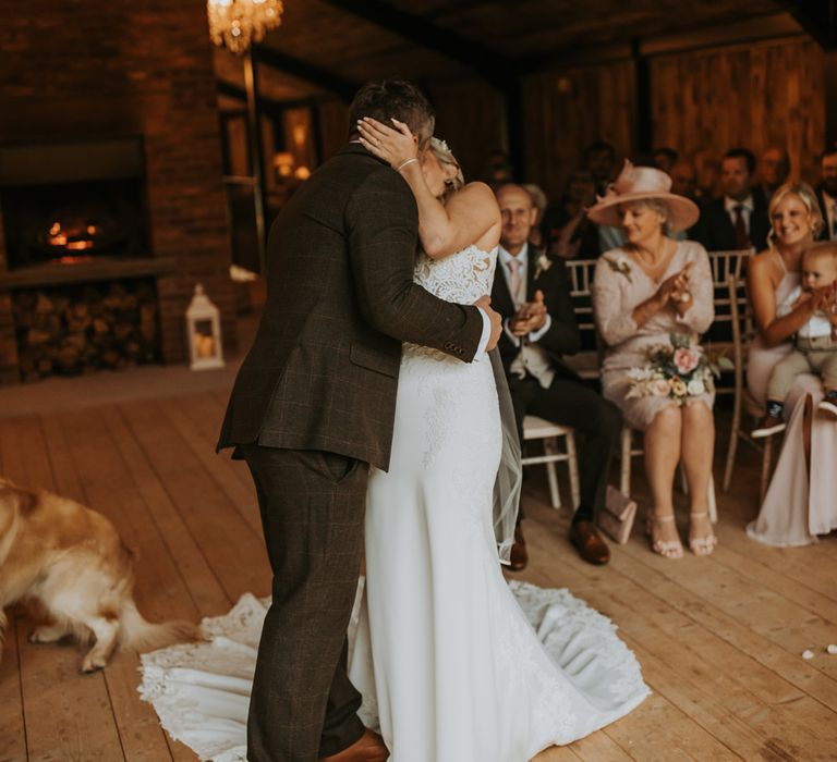 Bride in lace wedding dress with train kisses groom in dark brown suit during rustic wedding ceremony at the Hidden River Barn wedding venue