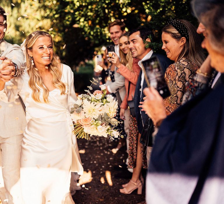 Smiling bride in satin dress and long sleeve wrap top holding pink, green and yellow bridal bouquet holds hands with groom in open white shirt and grey suit as guests throw petal confetti 