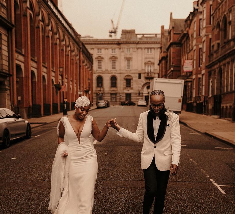 Black bride in a fitted Pronovias wedding dress with plunging neckline and beaded cap headdress holding hands with her groom in a white tuxedo jacket on the streets on Birmingham 