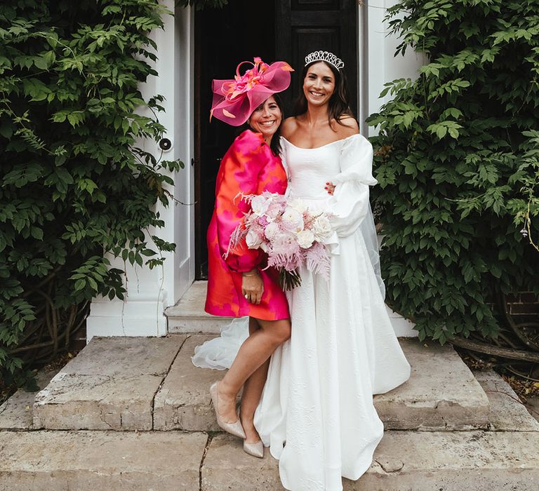 Bride in Emma Beaumont dress and tiara crown with pink and white wedding bouquet stands with guest in bright pink and orange dress