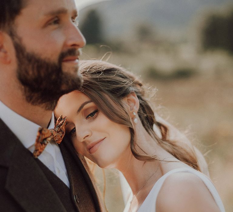 Bride wearing pearl drop earrings puts her head on the groom's shoulder who wears a orange paisley bow tie