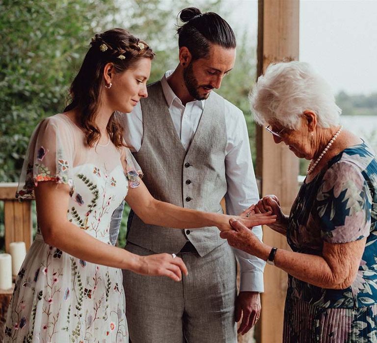 Wedding guest takes a closer look at the bride's wedding rings in floral embroidery wedding dress with groom in white shirt and grey waistcoat 