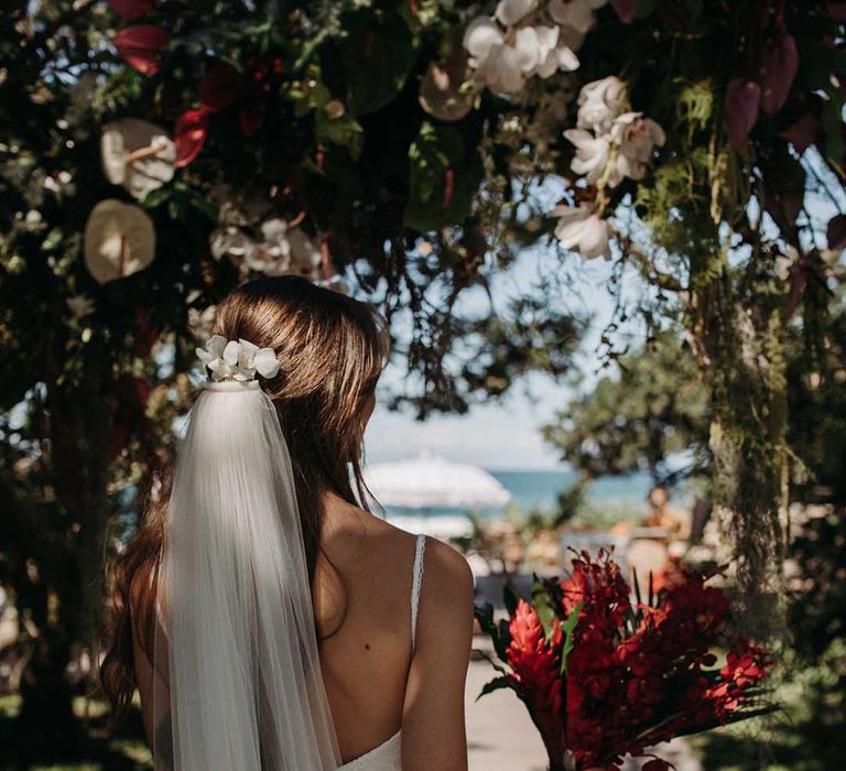 Bride in lace beach wedding dress and veil holding bright red flower wedding bouquet for beach wedding ceremony in South Africa 