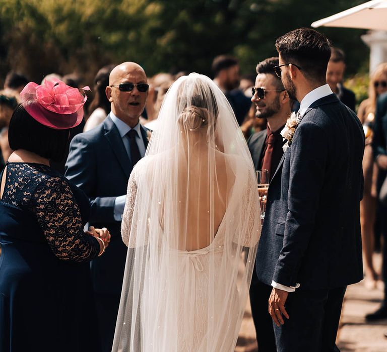 Bride in long sleeve beaded wedding dress with low back and pearl embellished veil socialises with wedding guests 