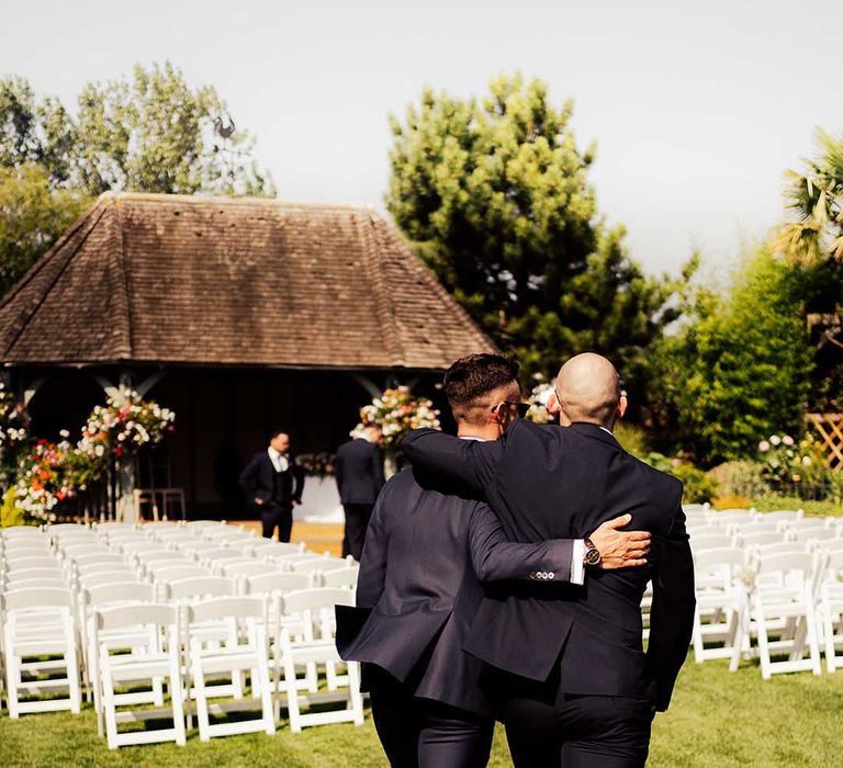 Groom walking into outdoor wedding ceremony area with best man as they have their arms around each other 