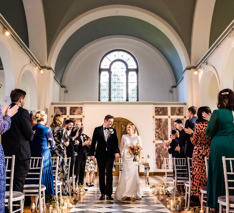 Groom in black tie and bride walk back down the aisle as a married couple with stained glass window