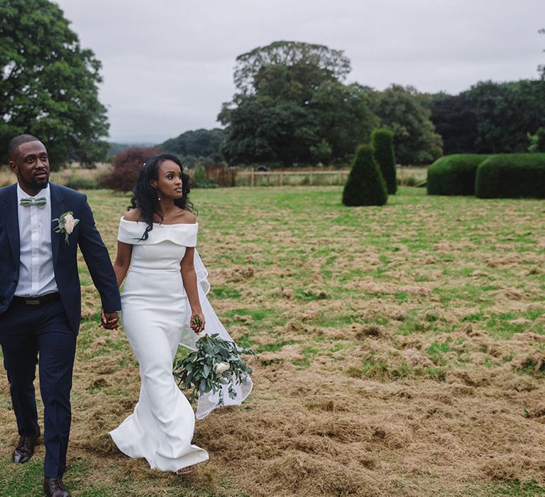 Bride in Bardot wedding dress and white flower bouquet with green leaves holding hands with groom in blue suit and mint green bow tie