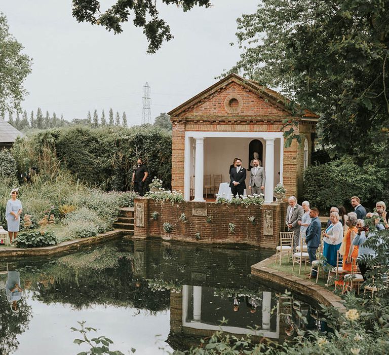 Wedding guests seated either side body of water as groom waits at the outdoor altar for the bride 