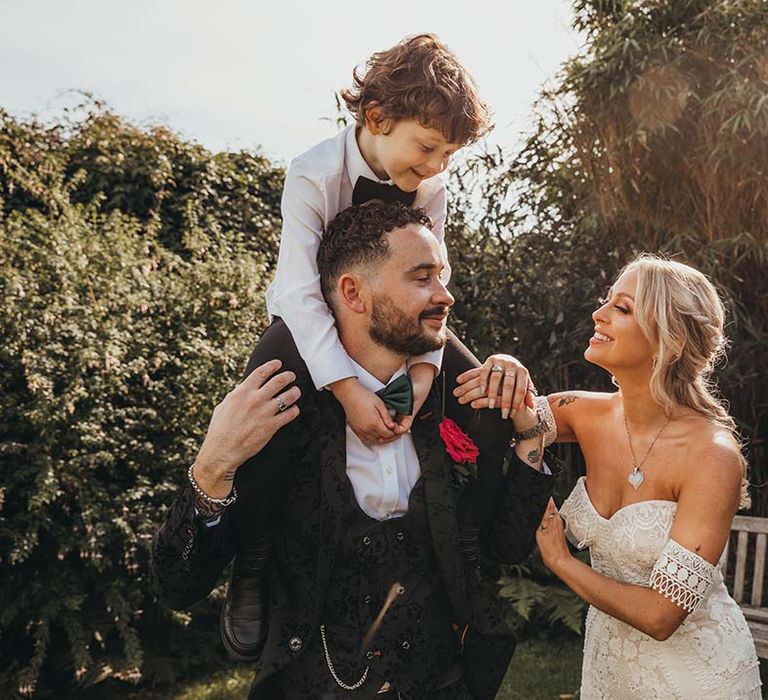 Bride in strapless boho lace gown with from in patterned tux with son on his shoulders in black tie