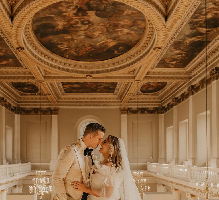 Bride and groom lean in for a kiss as they stand on balcony overlooking their reception with painted ceiling at The Banqueting House