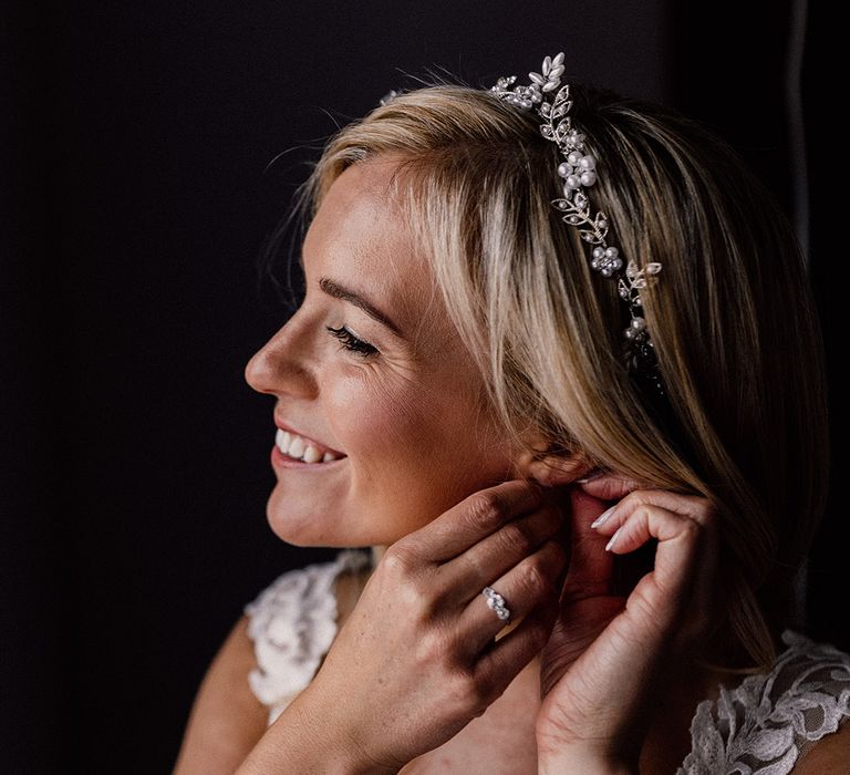 Blonde bride puts in her earrings wearing a gold leaf and pearl headband with a three stone engagement ring 