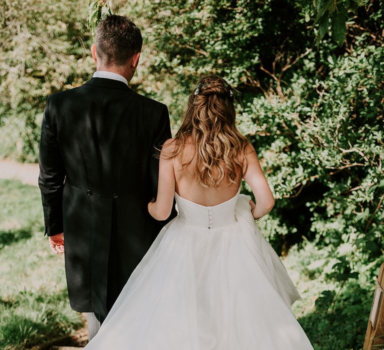 Bride wears her blonde hair in loose curls and low backed dress whilst walking alongside her groom 