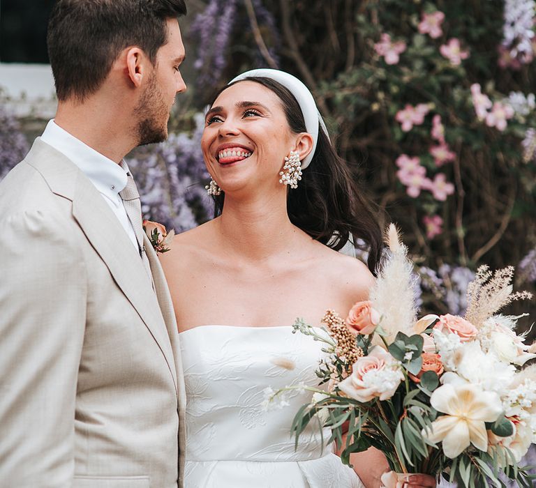 Bride wearing cool accessories smiles at the groom in a neutral suit 