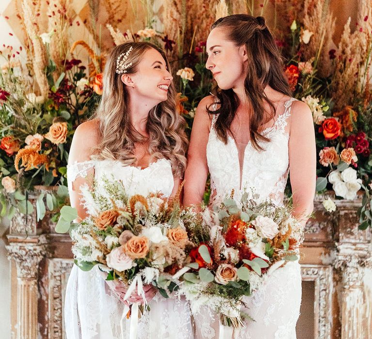 Brides look into each other's eyes in lace wedding dresses holding matching autumn wedding flower bouquet 