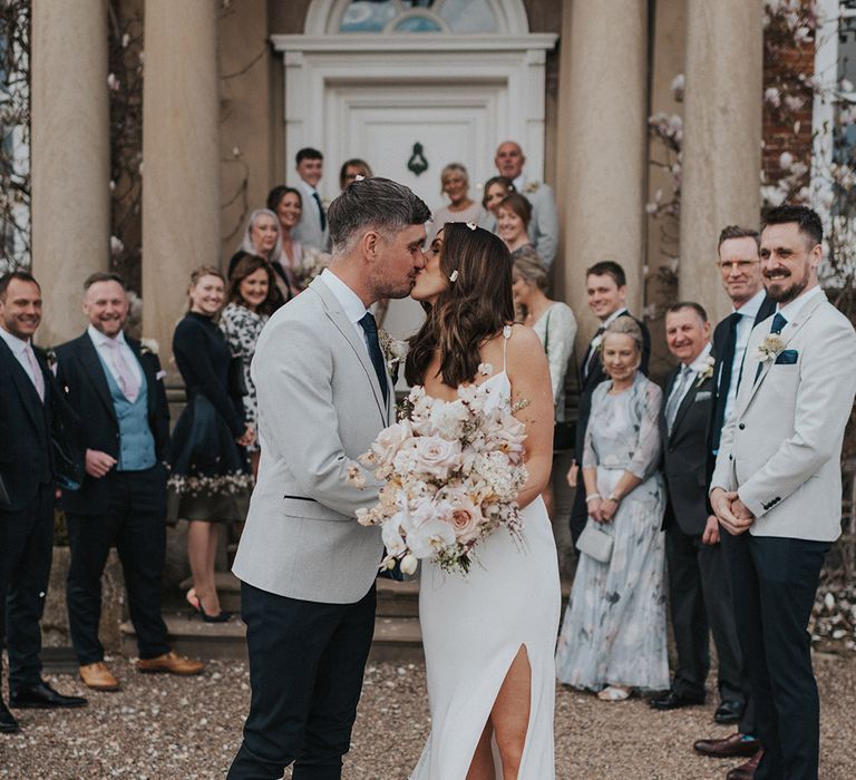 Bride in satin cowl neck wedding dress with split leg detail with groom in grey suit jacket and black trousers share a kiss 