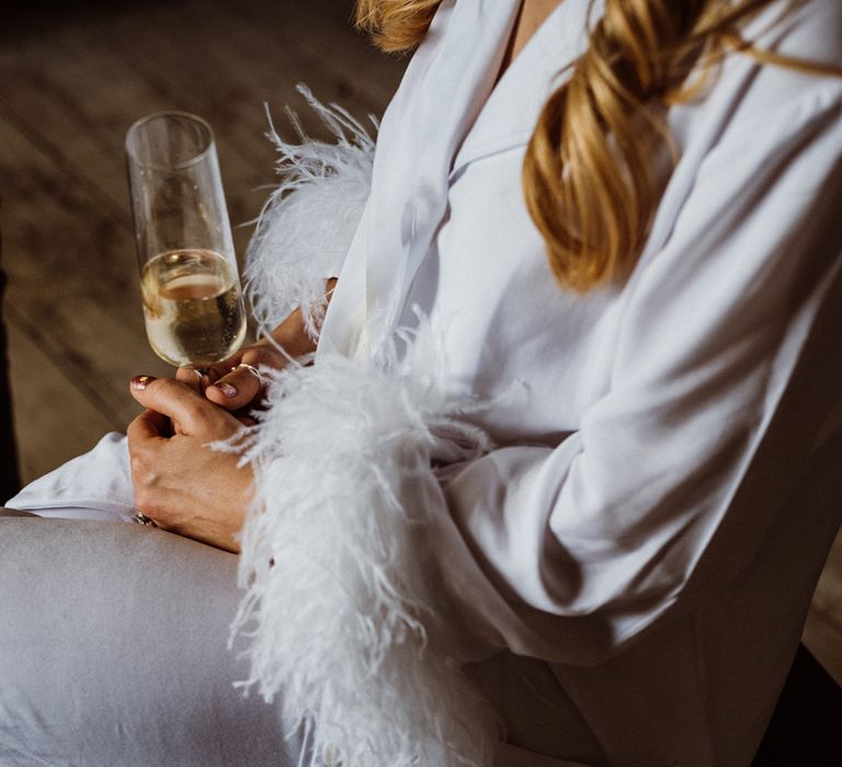 Bride holding a glass of champagne in white pyjamas with feather cuff 