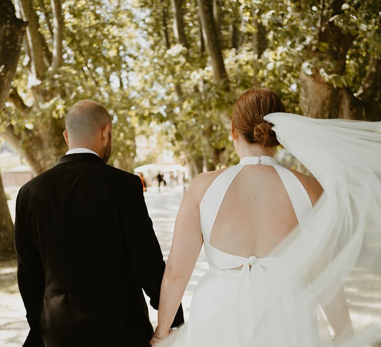 Brides veil blows in the wind and she walks with her groom wearing black tie after outdoor wedding ceremony in France 