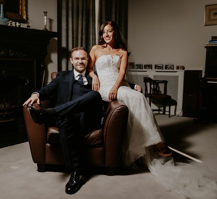 Bride wears Pronovias lace wedding dress and sits beside her groom in blue three piece suit on leather armchair 