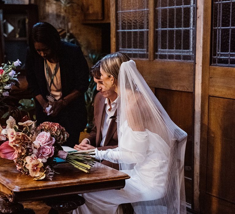 Bride wearing a pleated veil sits with the groom in a brown suit to sign the register 