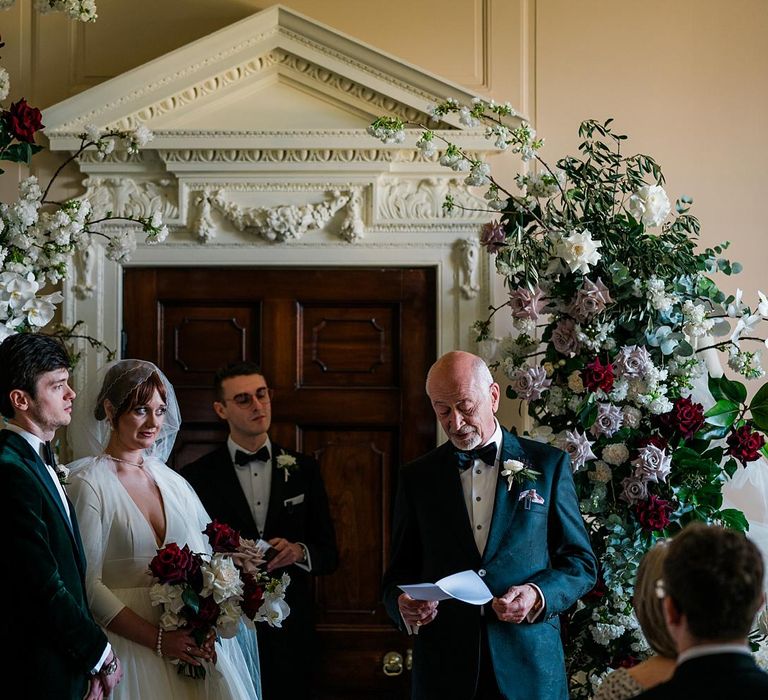 Bride & groom stand between floral archway at Findon Place during wedding ceremony 