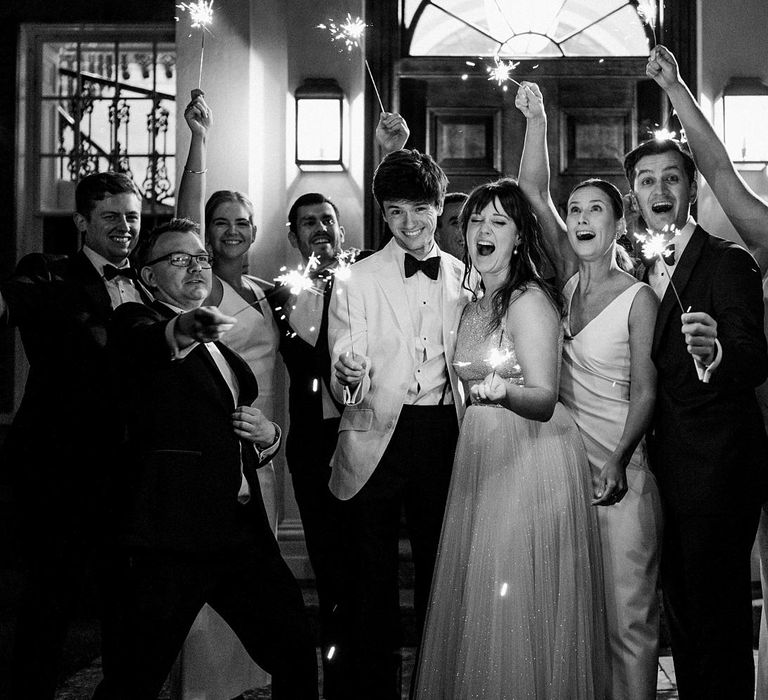 Black & white image of bride & groom holding sparklers with wedding guests outdoors 