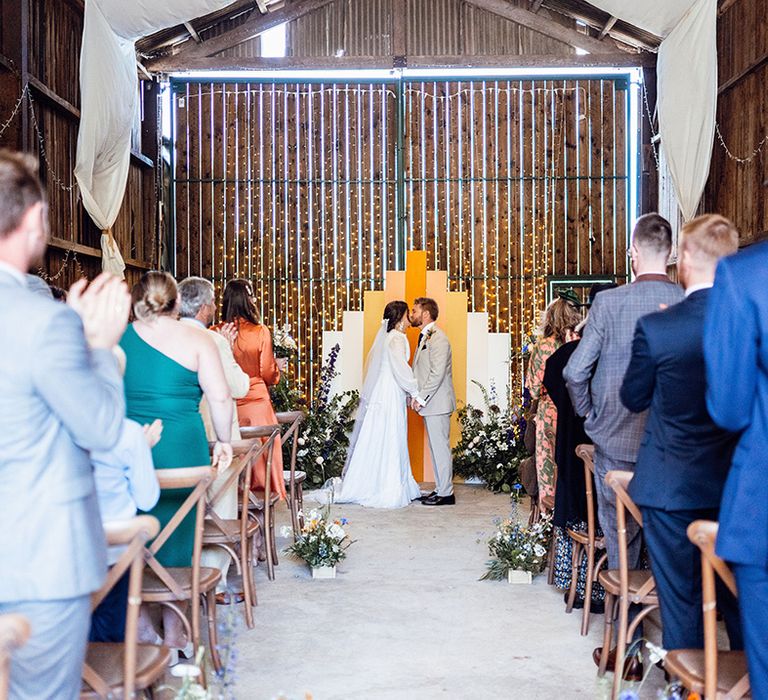 Bride and groom share their first kiss as a married couple in front go their orange, yellow and white wedding screen altar decor 