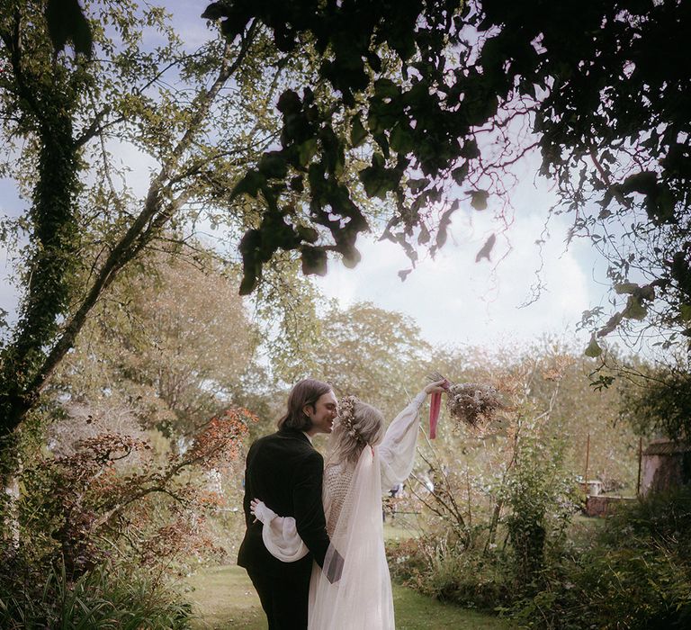 Bride wears dried floral flower crown with veil as she walks alongside her groom in green suit outdoors 