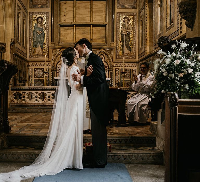 Bride in batwing sleeve and polka dot detail wedding dress and groom in black suit sharing a kiss at alter of church wedding with gold accents