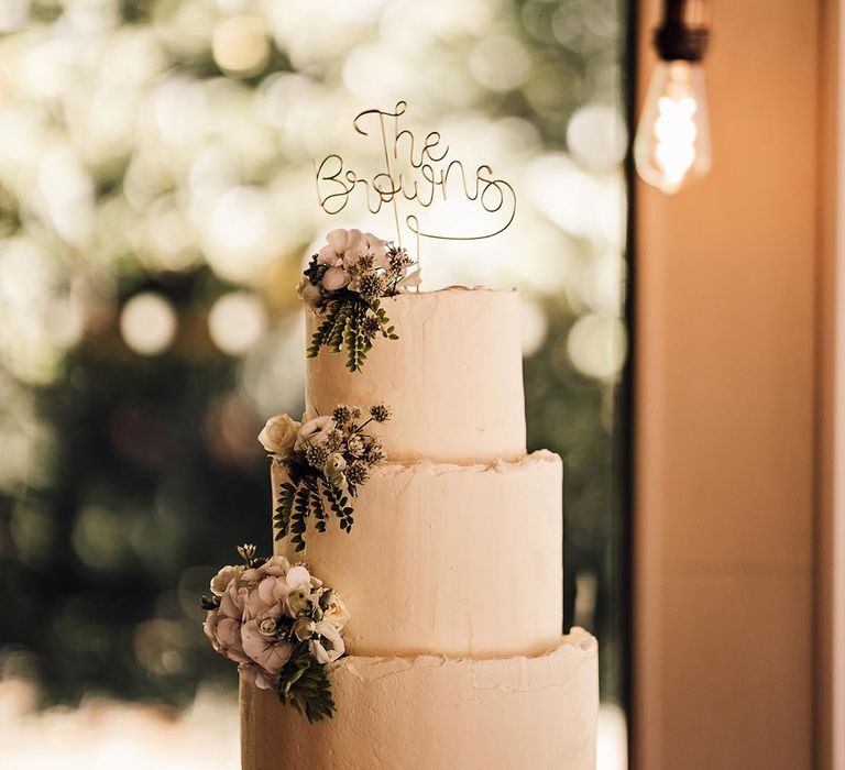 Three tier white frosted wedding cake with a personalised gold wire cake topper and decorative flowers