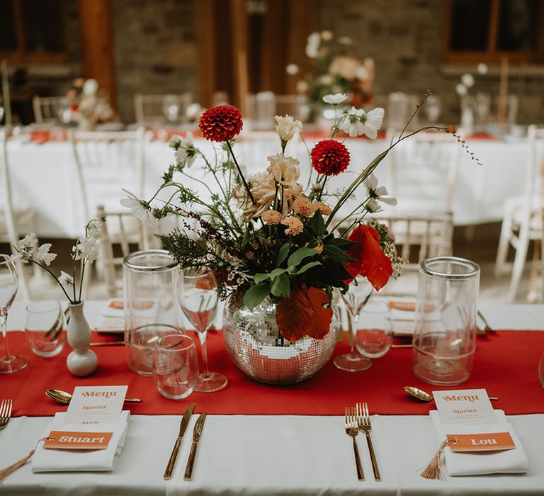 White table cloth with red table runner and disco ball vase holding a red and neutral floral arrangement with 70's-inspired wedding menu 