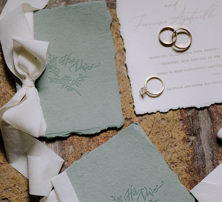 Sage green vow books tied with white ribbon bows with gold wedding rings and engagement ring resting on a white wedding invitation.