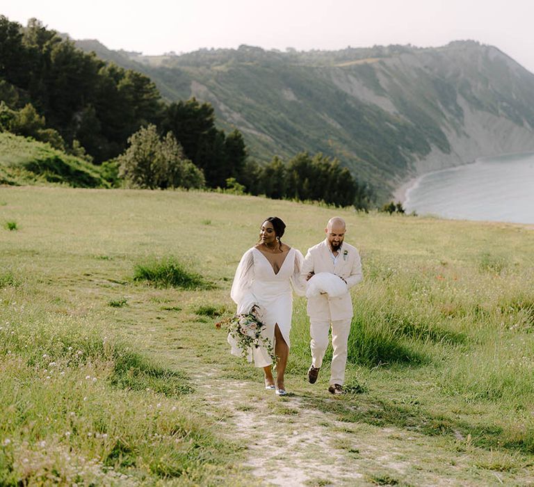 Groom holds Bride’s train as they walk through fields on the Italian coast