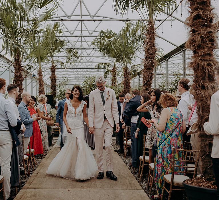 Bride and Groom walk up the palm tree lined aisle smiling and holding hands