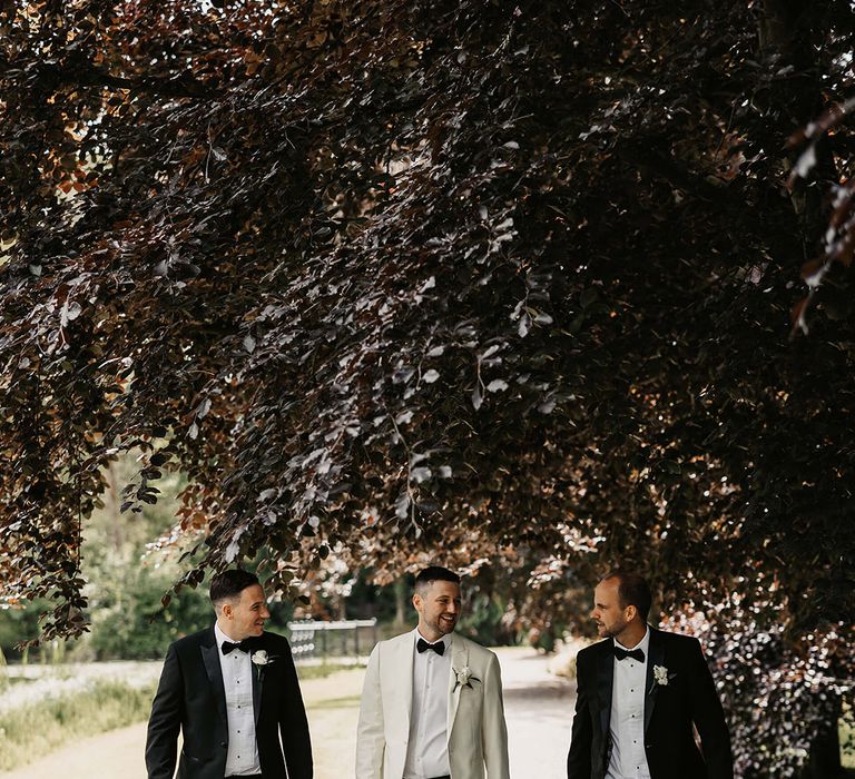 The groom in a white tuxedo walks with two of the groomsmen in black tie all wearing white rose buttonholes 