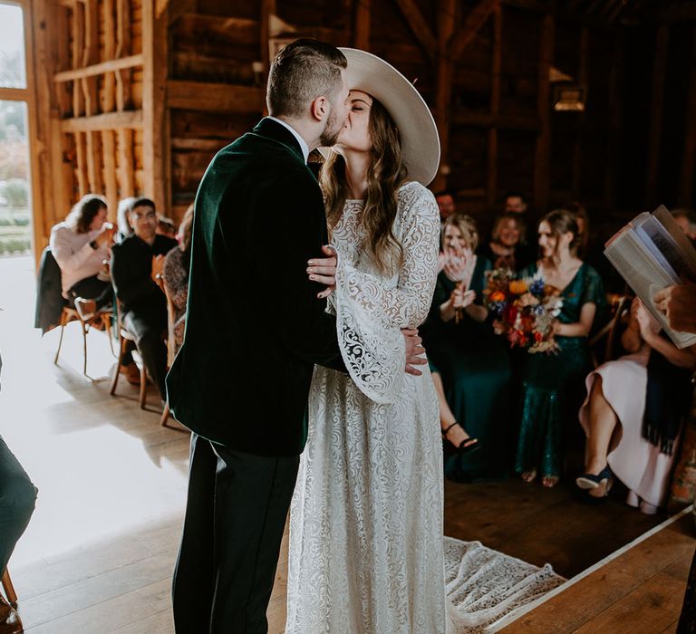 The groom in a green velvet suit jacket kisses the bride in a boho lace wedding dress and hat with dried flowers 