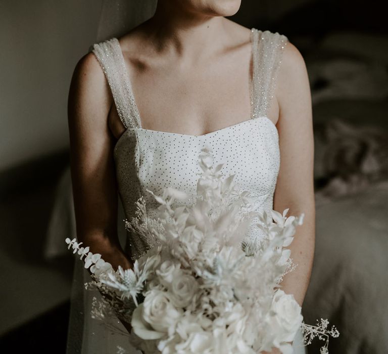 Bride in sparkly wedding dress and straps holding all white bouquet with dried flowers and roses wearing silver pearl earrings 