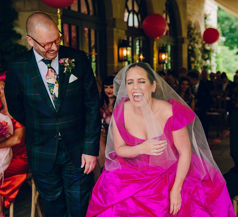 Groom in dark forest green tartan suit with patterned tie, white pocket square and pink and green boutonniere and bride laughing in off the shoulder front ruching pink wedding dress and two layer white and pink ombre veil 