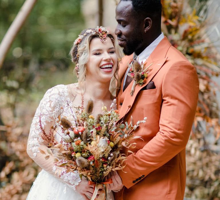 Groom in burnt orange rust coloured suit with brown tie, pocket square, brown accessories and dried flower, rose and pampas grass boutonniere and bride in long sleeved v neck soft ivory dress with lace floral detailing on the bodice and sheer sleeves holding large autumnal dried flower bouquet with red roses, foliage and pampas grass