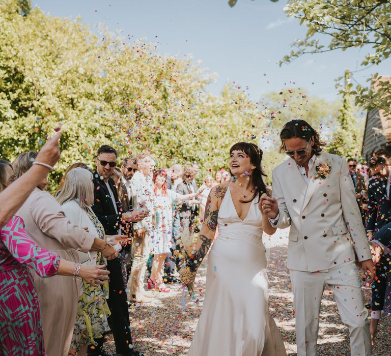 Bride in silk-look v-neck sleeveless ASOS wedding dress and groom in neutral coloured double-breasted ASOS suit and dried flower boutonniere doing confetti walk after wedding