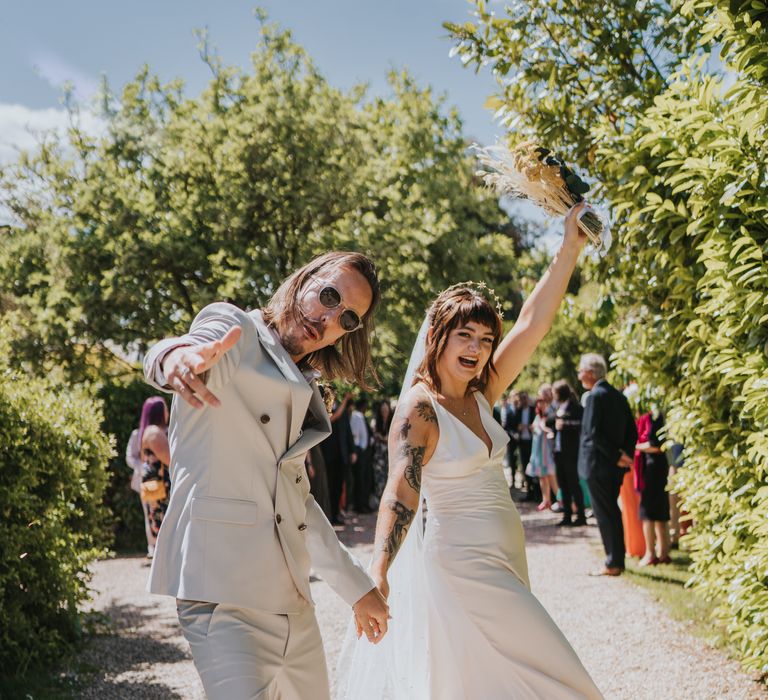 Groom in neutral coloured double-breasted ASOS suit and black high-top converse with dried flower and foliage boutonniere and bride in silk-look v-neck sleeveless ASOS wedding dress holding hands and posing at The Reid Rooms wedding venue